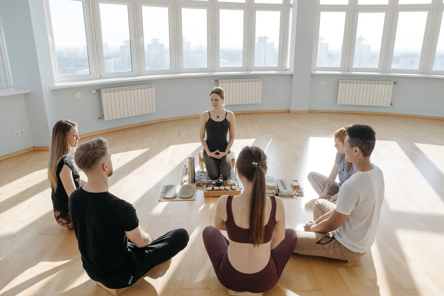 Five people sitting cross-legged in a circle on a sunlit wooden floor, facing a woman who kneels behind a tray of tea cups.