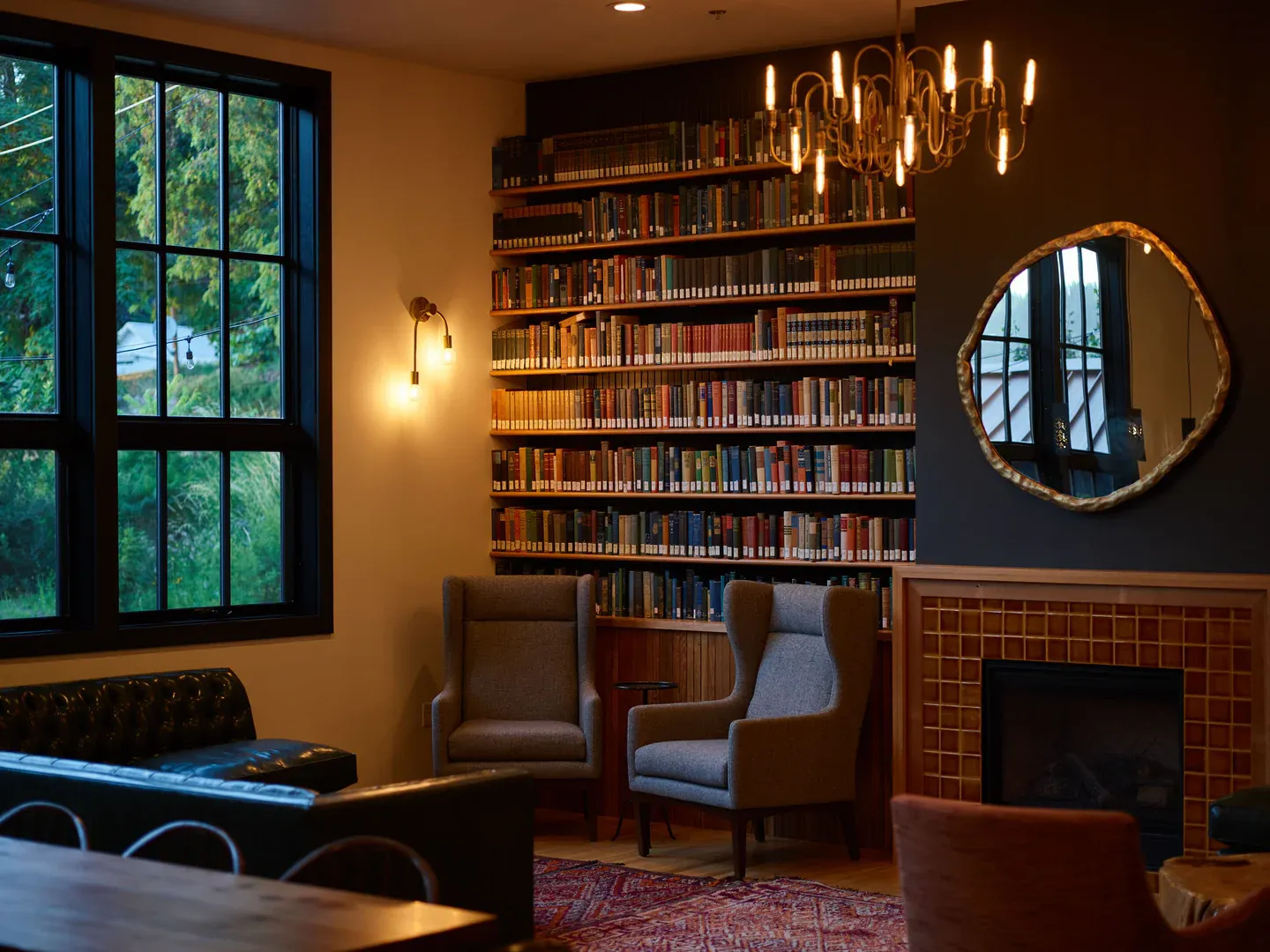 Wingback armchairs beside a fireplace in a room lined with bookshelves, with trees outside the tall windows.